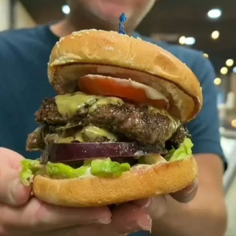 A person holds a large Secret Sammich burger with two patties, cheese, lettuce, onion, and tomato.
