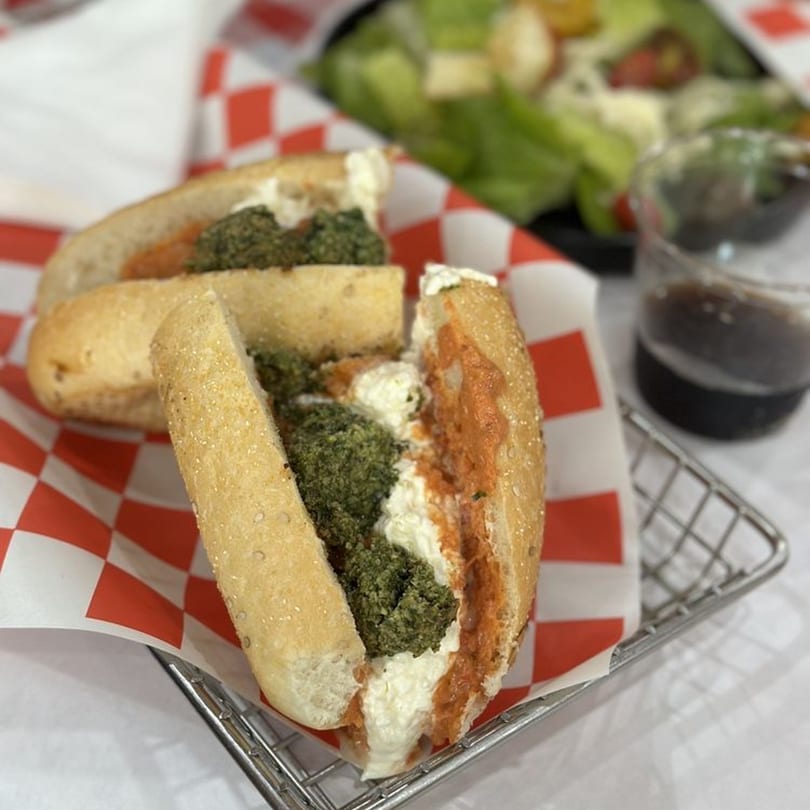 Two Secret Sammich deli sandwiches with pesto, red sauce, and cheese in a metal basket.
